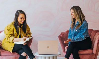 Two women sitting in pink chairs, one journaling and the other gesturing while talking, a laptop on a small table between them.