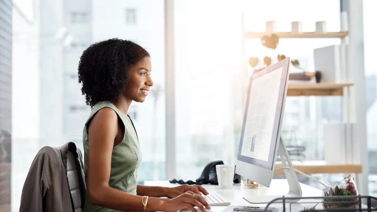 Woman in a green sleeveless top with curly hair smiles while typing on a computer at her desk in a bright office.