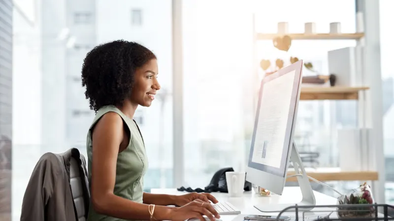Woman in a green sleeveless top with curly hair smiles while typing on a computer at her desk in a bright office.