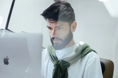 Man with dark hair and a beard focused on a computer screen, wearing a white t-shirt with a green sweater tied around his neck.