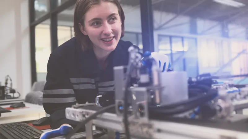 A young female engineer in a dark uniform with reflective stripes works intently on a complex piece of machinery.