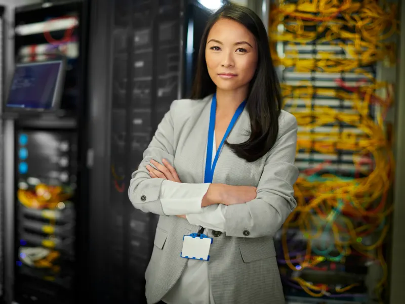 Woman in a light gray blazer stands with arms crossed in front of server racks filled with colorful cables.