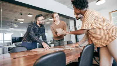 Three people gathered around a wooden conference table looking at documents; one woman holds a paper and gestures.