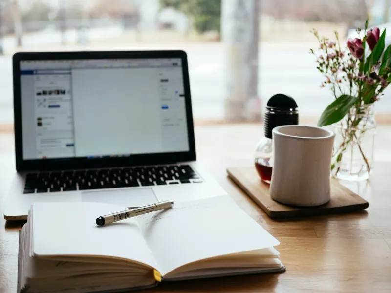 Laptop, open notebook, pen, mug, and flowers on a wooden table, suggesting a workspace or cafe setting.