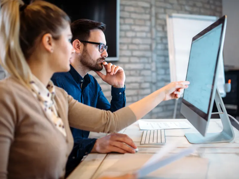 A woman points to a computer screen as a man looks on thoughtfully, working together in an office.