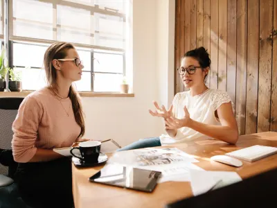 Two women in glasses having a meeting at a wooden desk with papers and a computer.