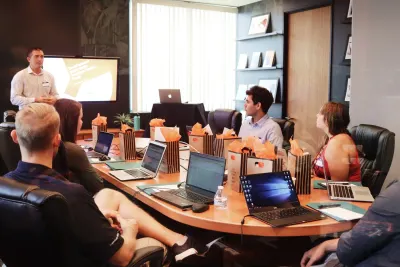 A presenter stands by a screen as a team sits around a conference table with laptops and gift bags.