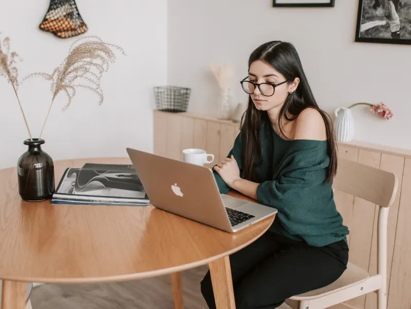 Woman with glasses in a green off-the-shoulder top working on a laptop at a wooden desk.