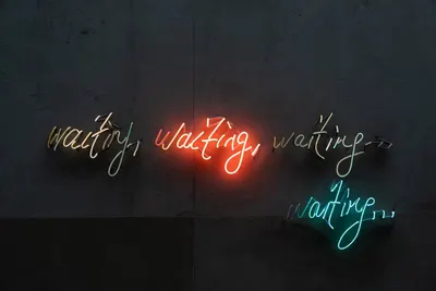 Four neon signs spell out the word “waiting” in yellow, red, white, and turquoise.