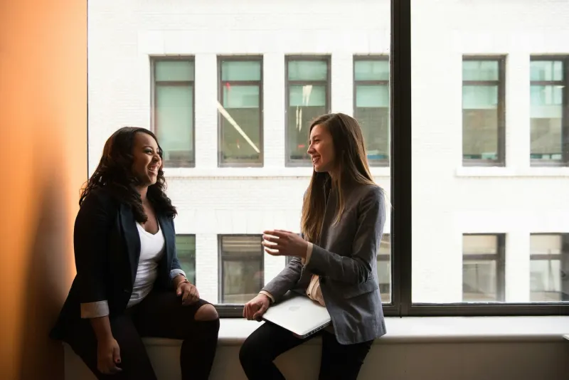 Two women in business attire laughing while sitting by a window with a laptop.