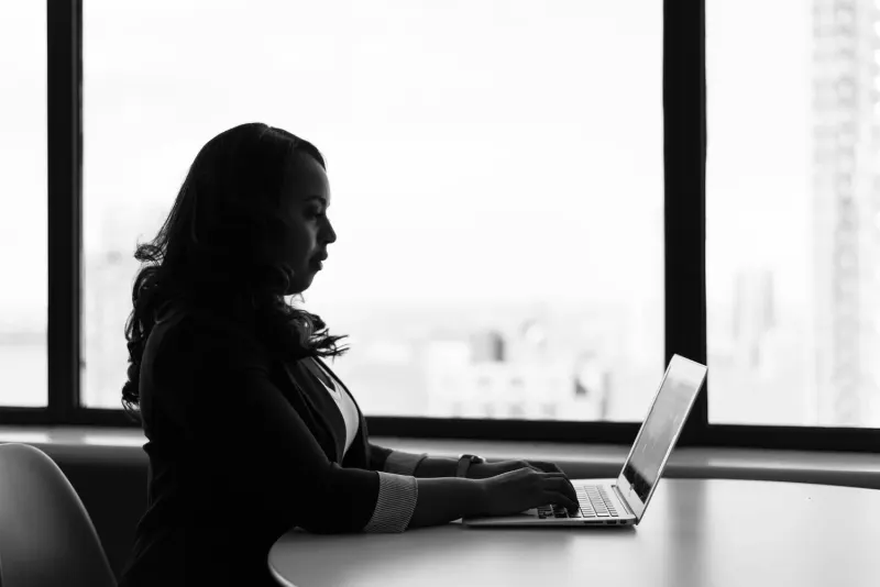 Silhouette of a woman in a blazer typing on a laptop in front of a large window with a city view.