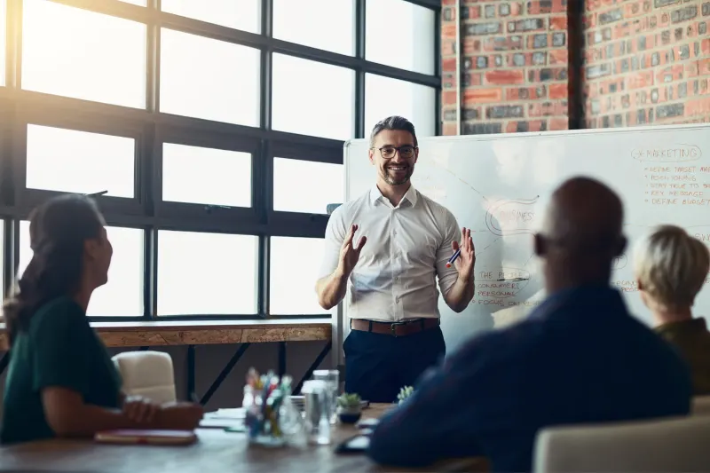 Smiling presenter at a whiteboard discussing marketing strategy with colleagues in a meeting room.