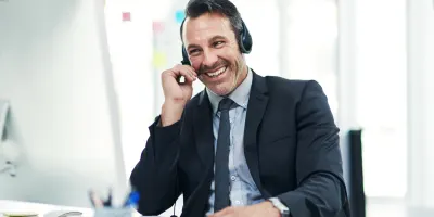 A smiling man in a suit wearing a headset answers the phone at his desk.