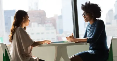 Two women sit at a table talking; one has long dark hair in a blush top, the other has dark curly hair and glasses.