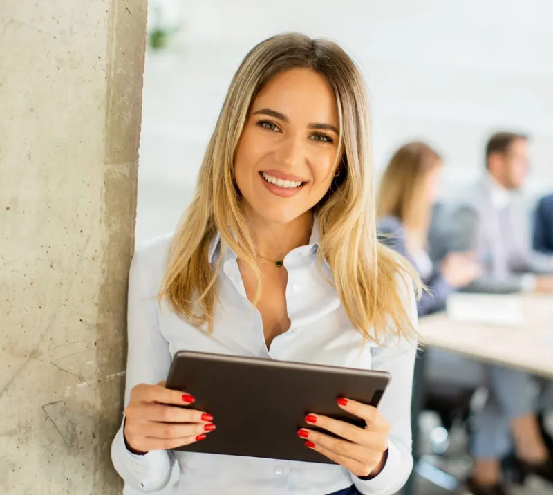 A smiling operations leader holding a tablet, leaning against a concrete pillar while her team meets at a table behind her