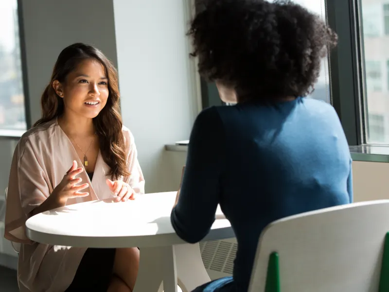 Two women have a discussion across a white table; one facing the camera gestures as she speaks.