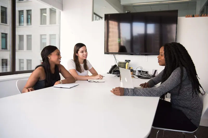 Three women at a conference table in a modern office; the woman on the left smiles toward the center.