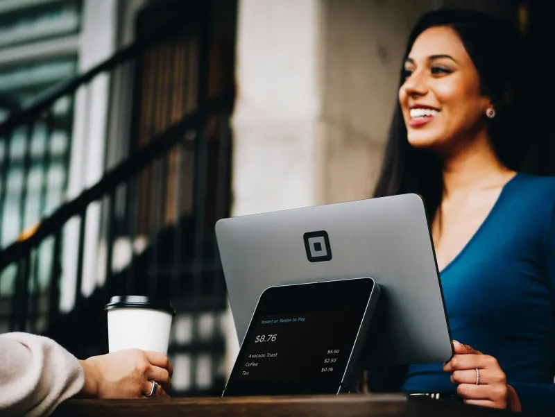 A smiling woman in a blue dress uses a Square Point of Sale system to process a payment of $8.76 for avocado toast and coffee.