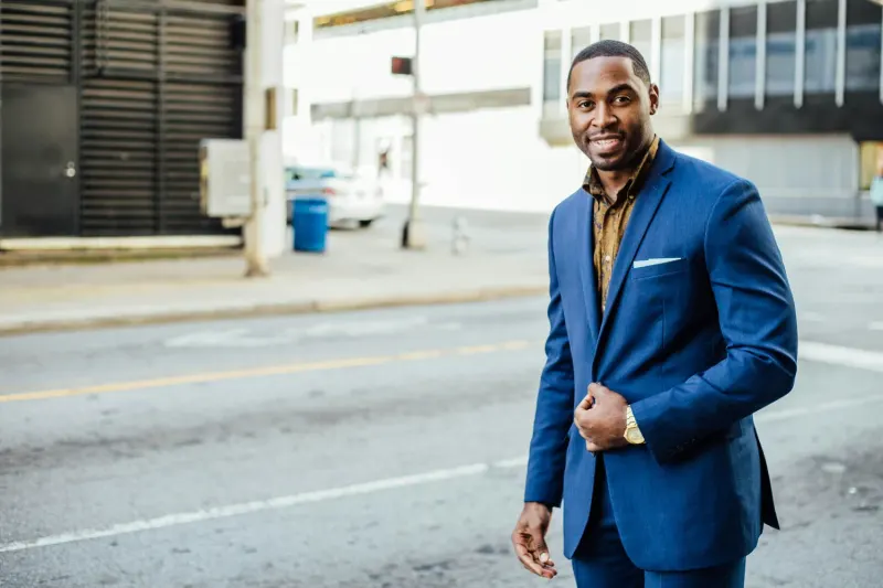 Man in a blue suit stands on a city street, smiling and holding the lapel of his jacket.