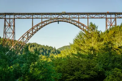 A large, rust-colored metal arch bridge spans a forested valley under a clear blue sky.