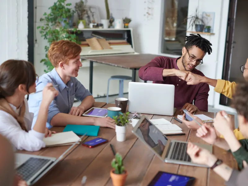 A diverse group of people collaborate around a wooden table sharing laptops, tablets, and notebooks, celebrating with a fist bump.
