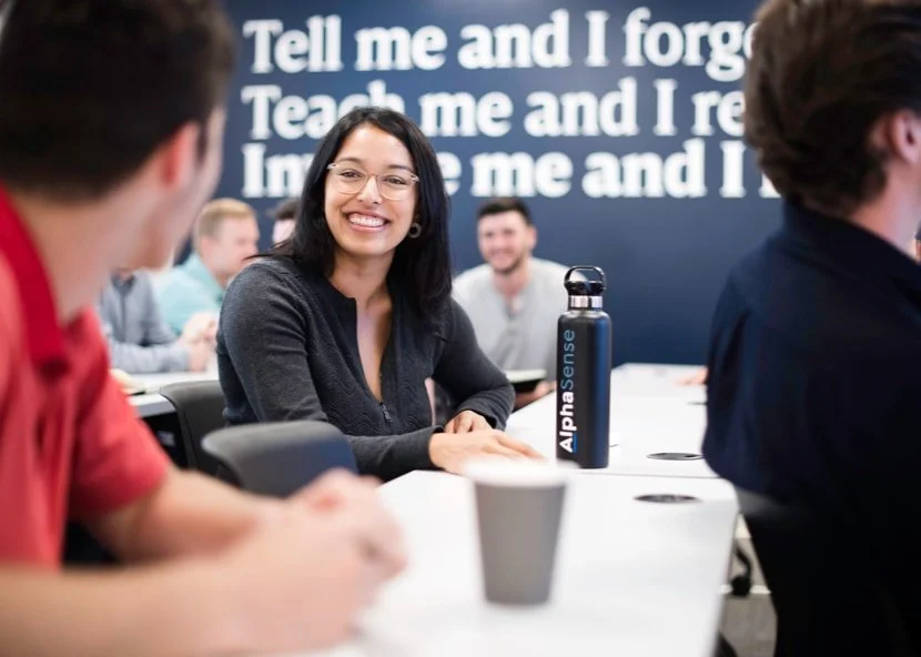 A smiling woman in glasses looks at someone across a table in a classroom setting, with an AlphaSense water bottle on the table.