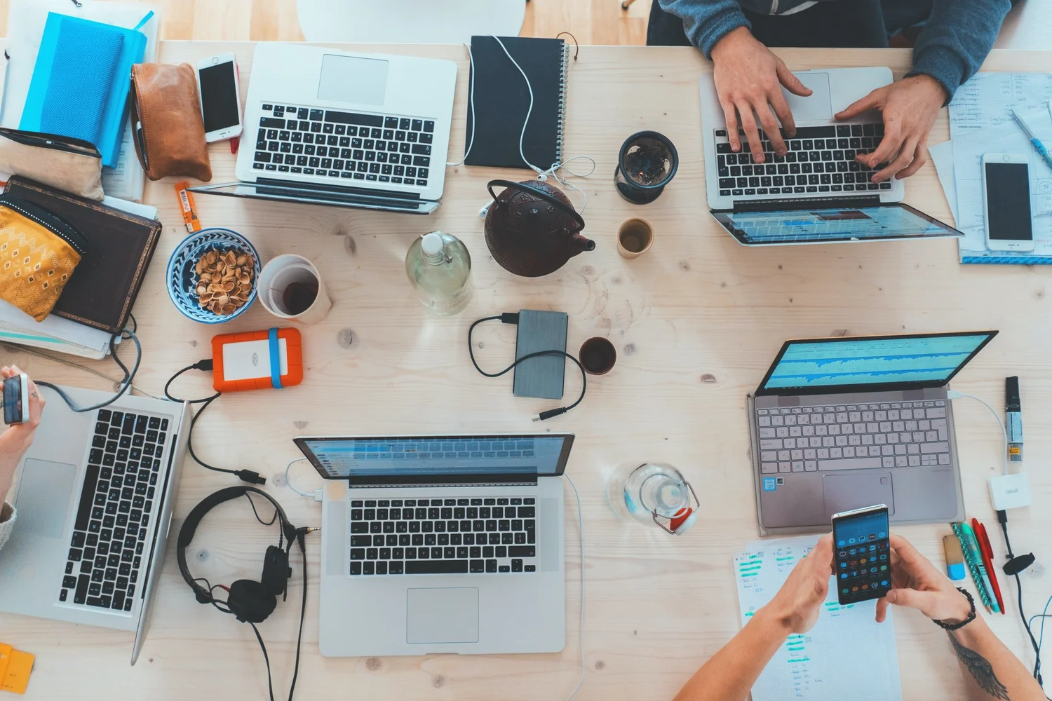 Overhead view of a wooden desk with multiple laptops, phones, headphones, and snacks, suggesting a collaborative workspace.