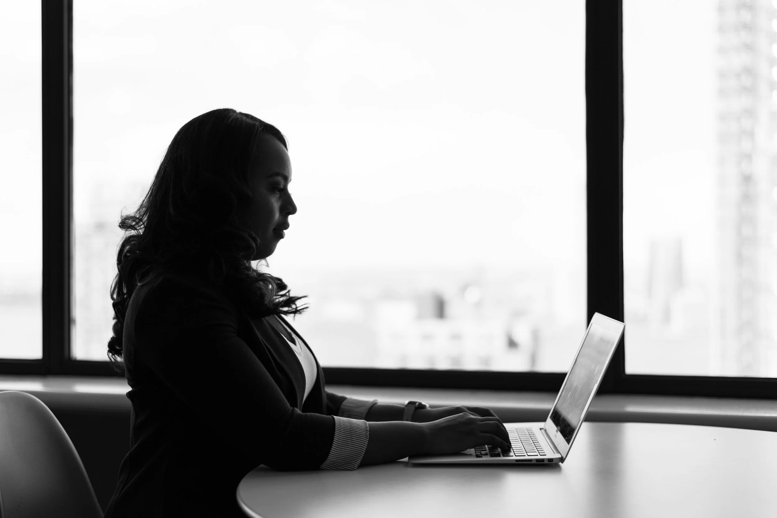 Silhouette of a woman in a blazer typing on a laptop at a table by a large window.