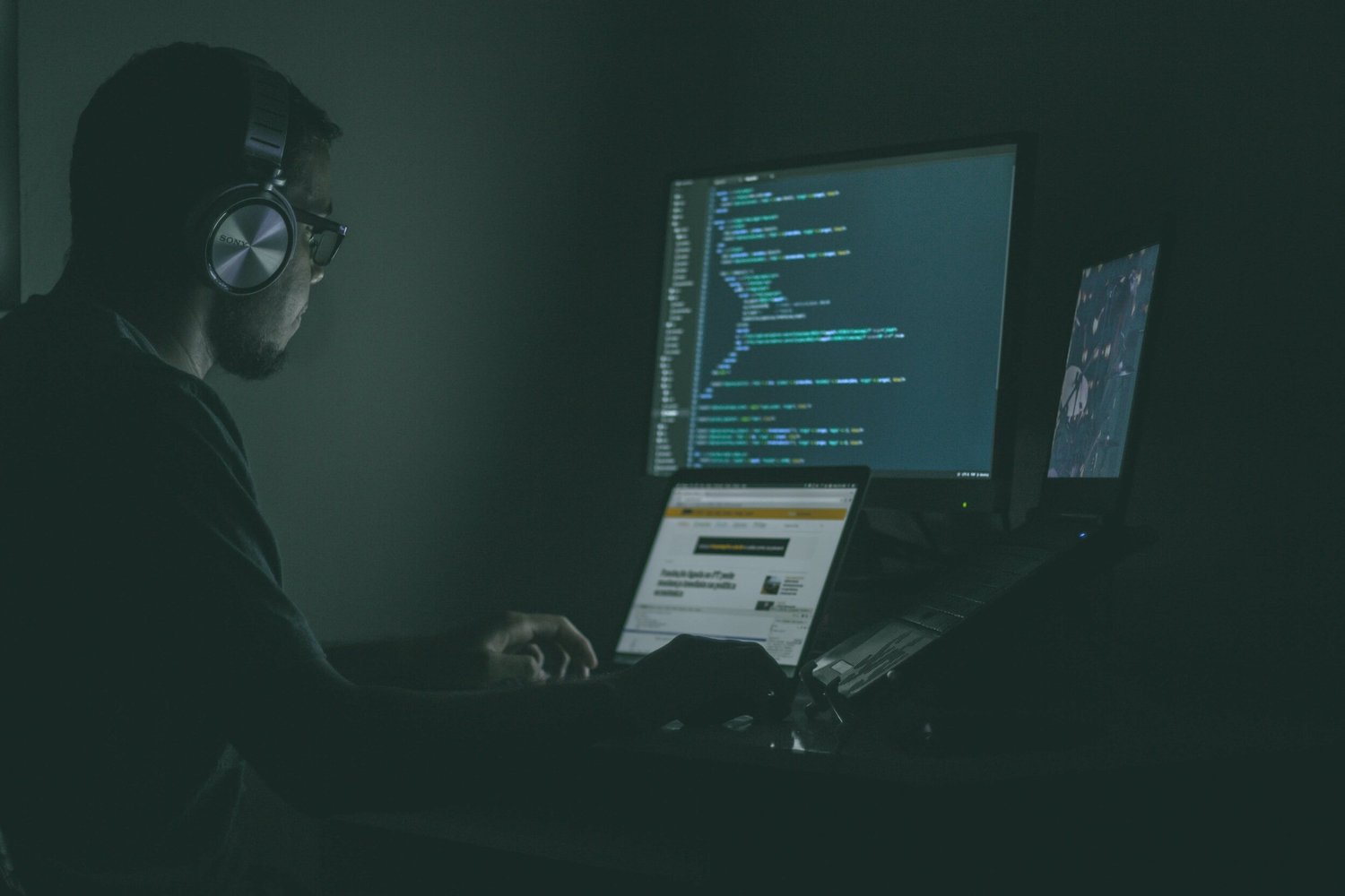Man wearing headphones and glasses types on a laptop in a dimly lit room, code displayed on multiple monitors.