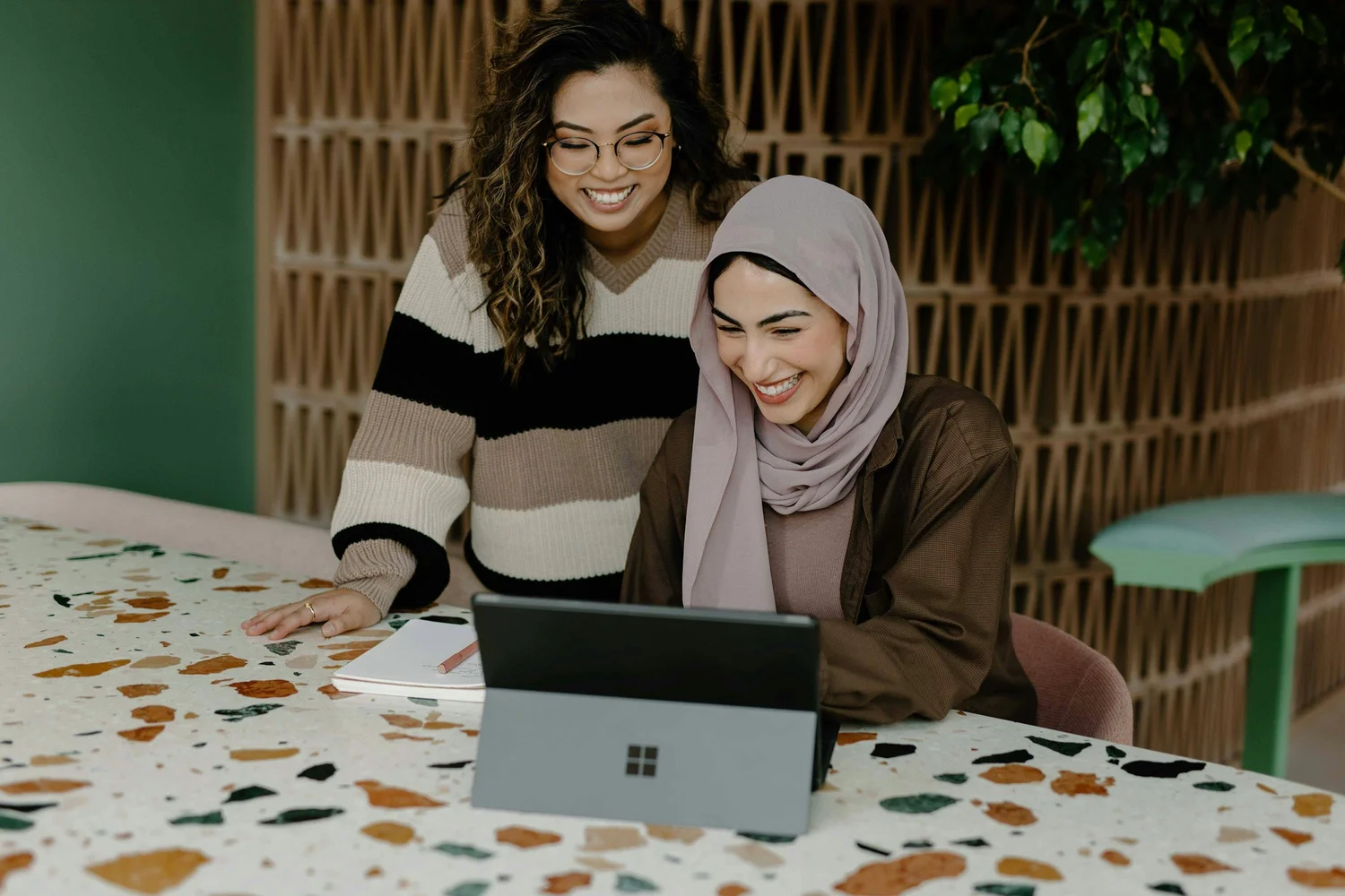 Two women of color laugh while looking at a laptop on a terrazzo table; one wears glasses and a striped sweater, the other a hijab.