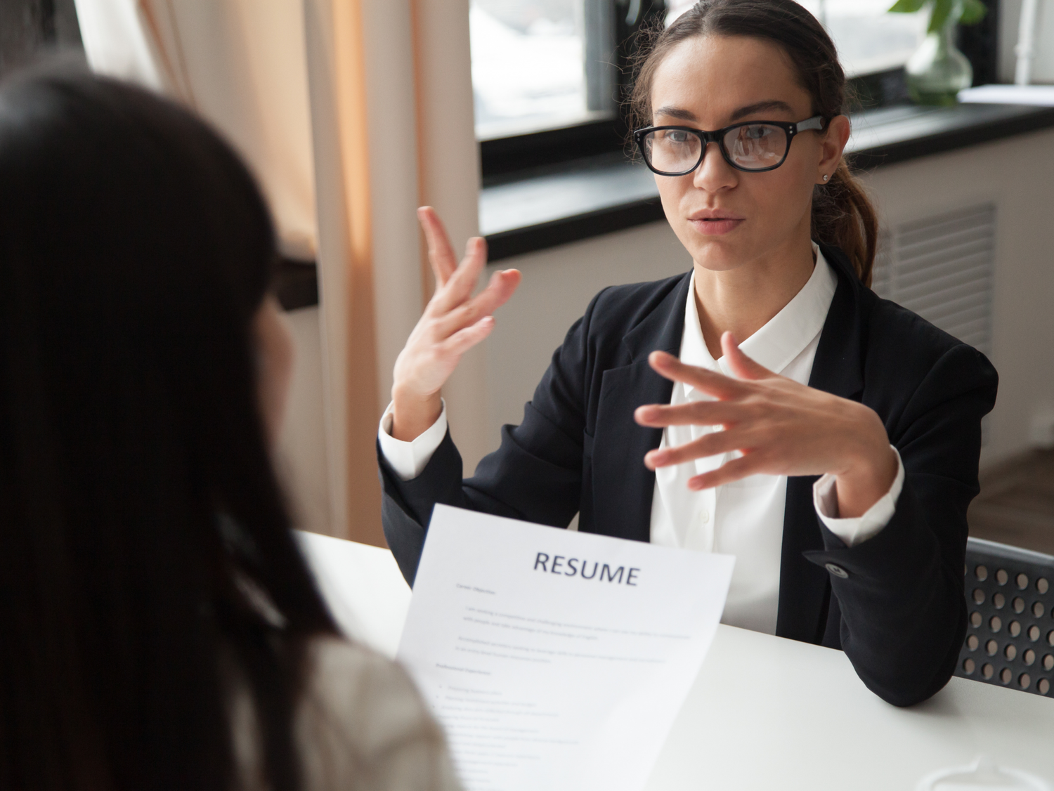 A woman in glasses and a black blazer interviews a candidate, gesturing with her hands as a resume sits on the table.