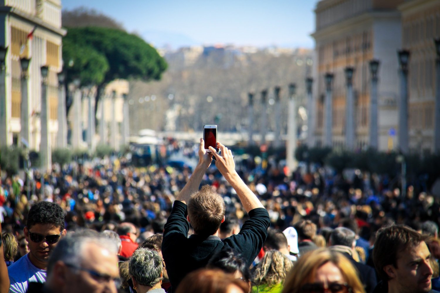 Back of a man’s head as he holds a phone up to record a large crowd gathered in a city square.