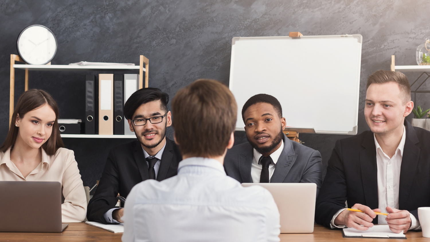 Four people sit at a table during a job interview, one facing away being interviewed.