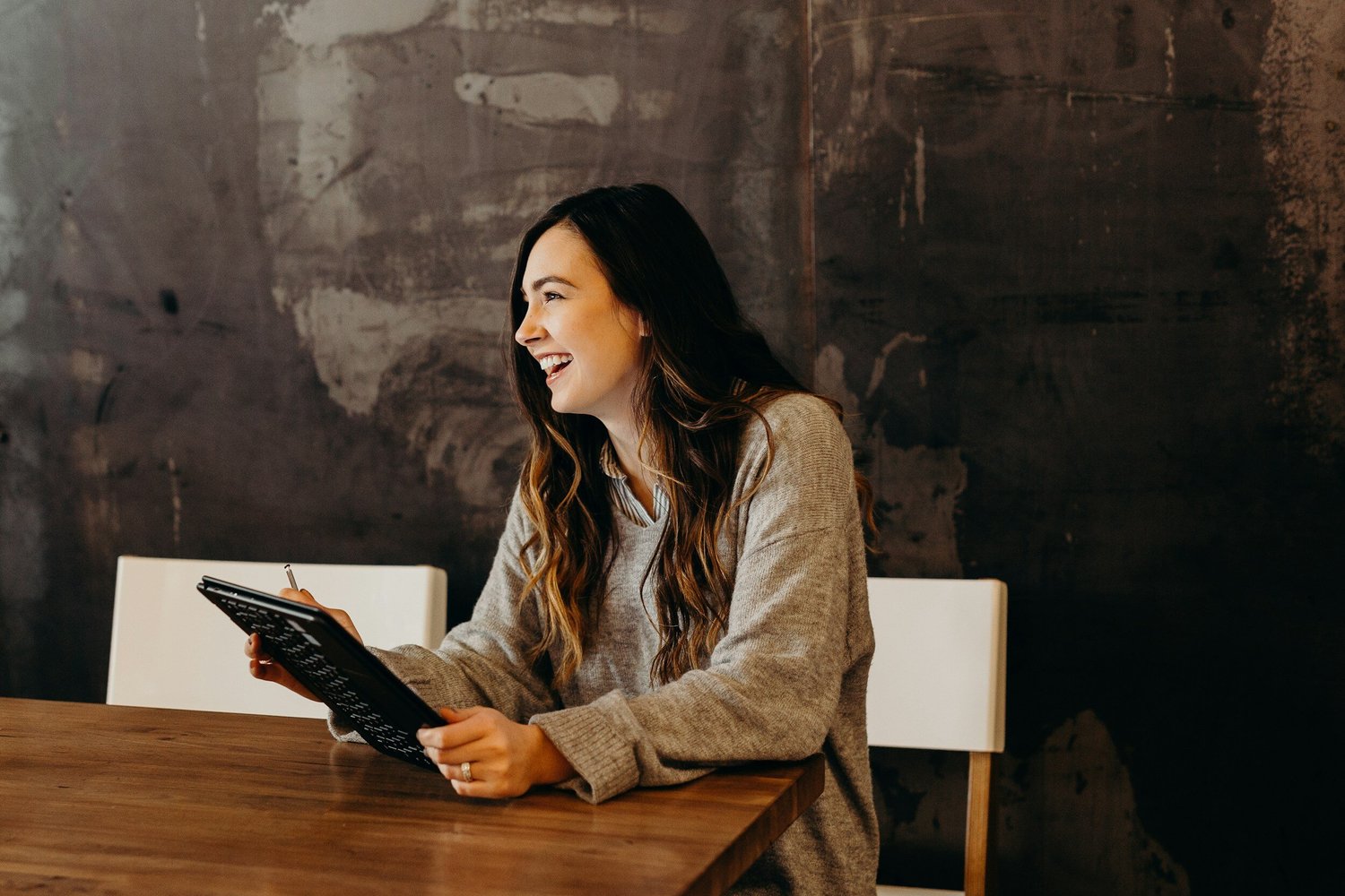 A woman with long wavy brown hair in a gray sweater laughs while holding a tablet and stylus at a wooden table.