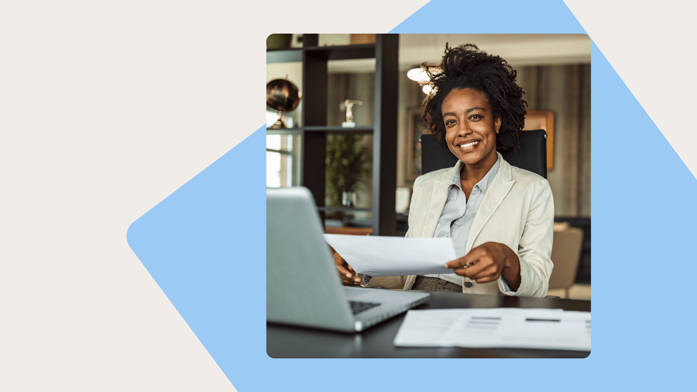 Smiling Black woman in a light blazer sits at a desk with a laptop and papers, holding a document.
