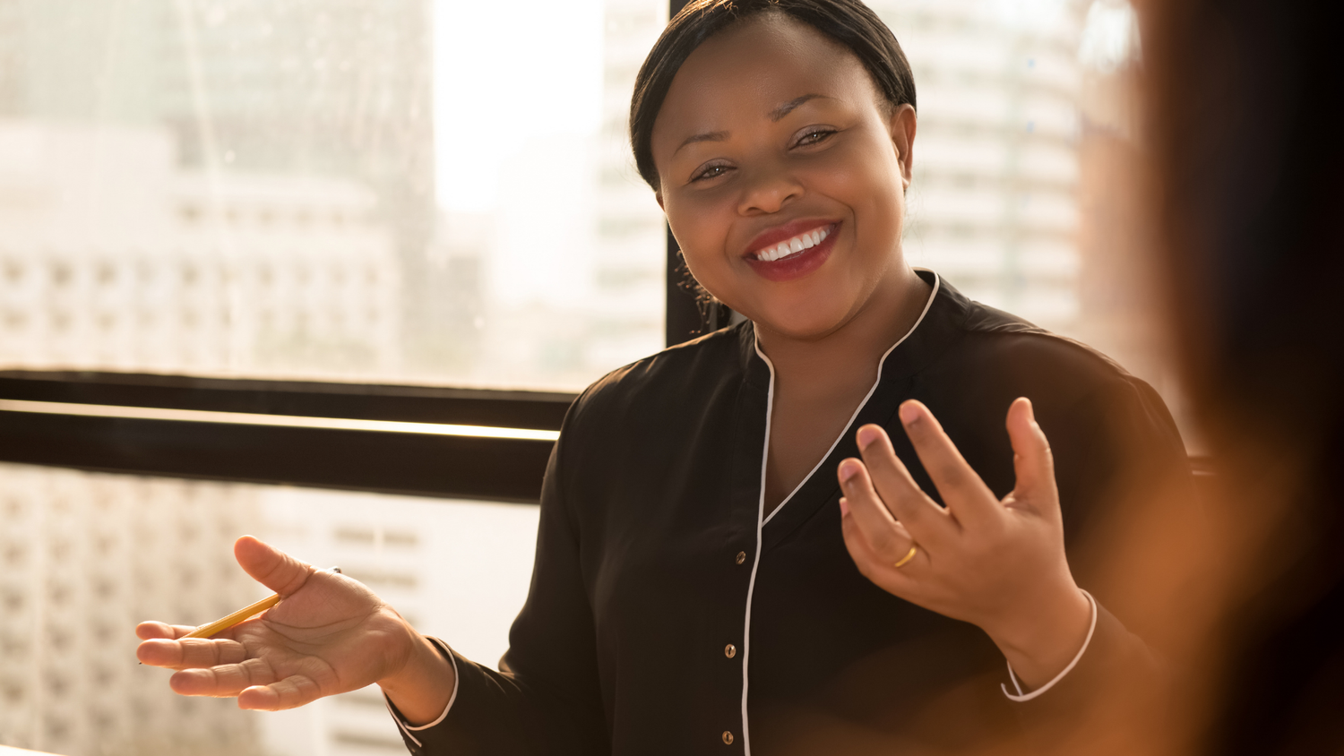 A woman in a black top smiles and gestures with her hands, holding a pencil, talking to someone out of frame.