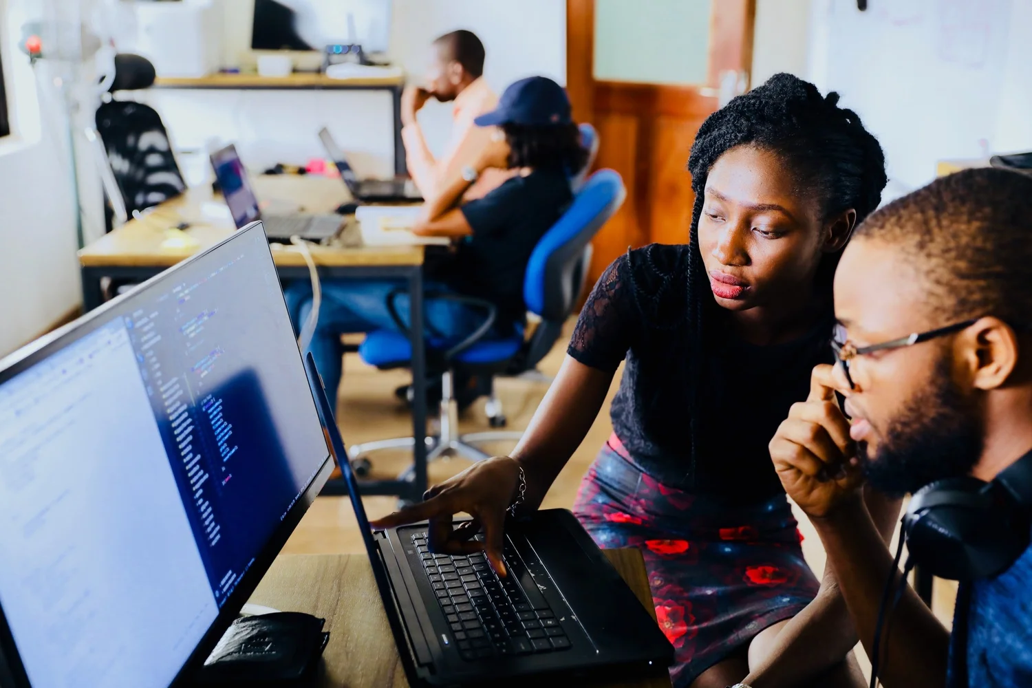 A Black woman and man collaborate over a laptop displaying code, another person working in the background.