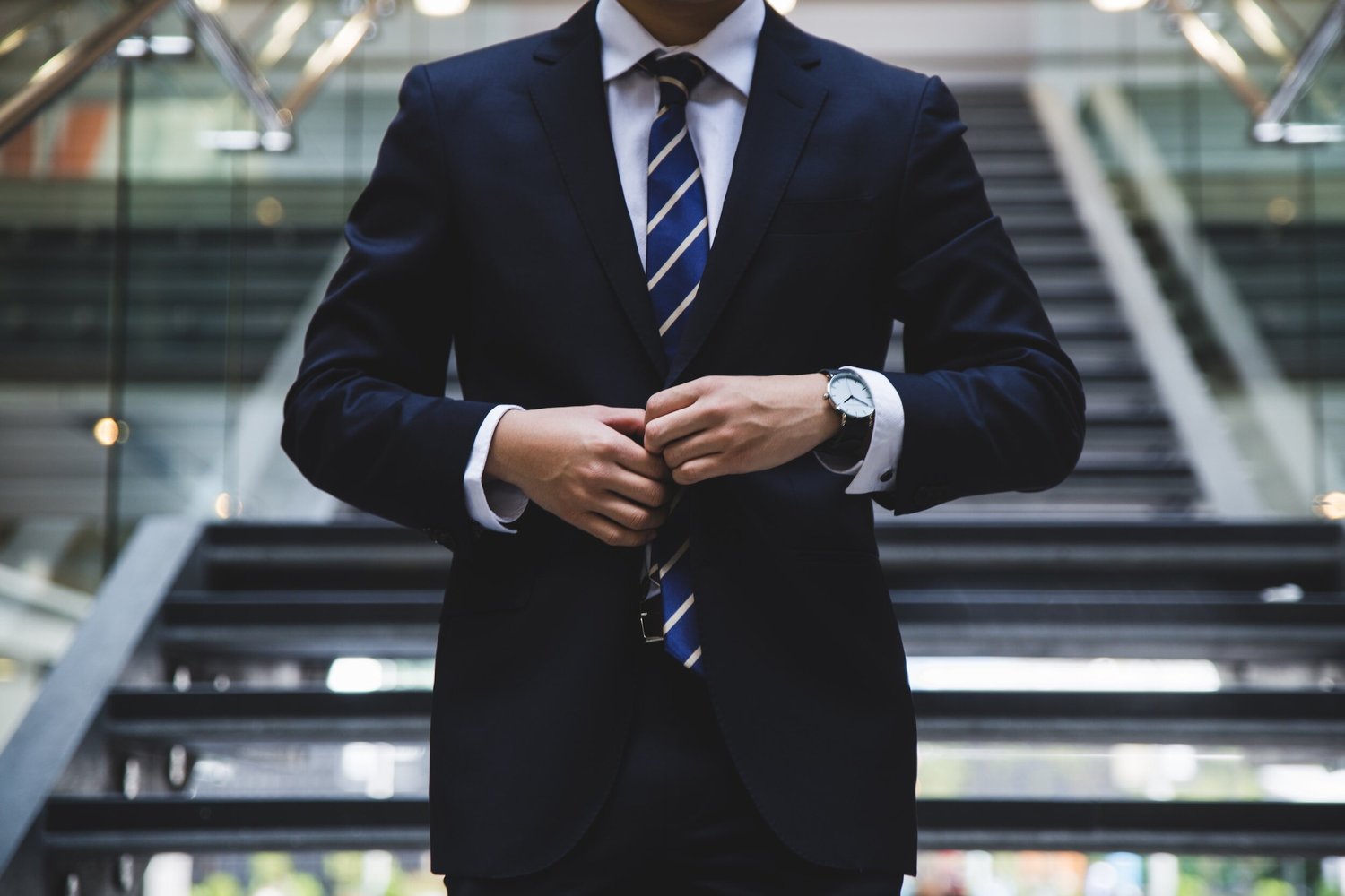 A man in a dark suit and striped tie fastens his jacket buttons while standing on stairs.