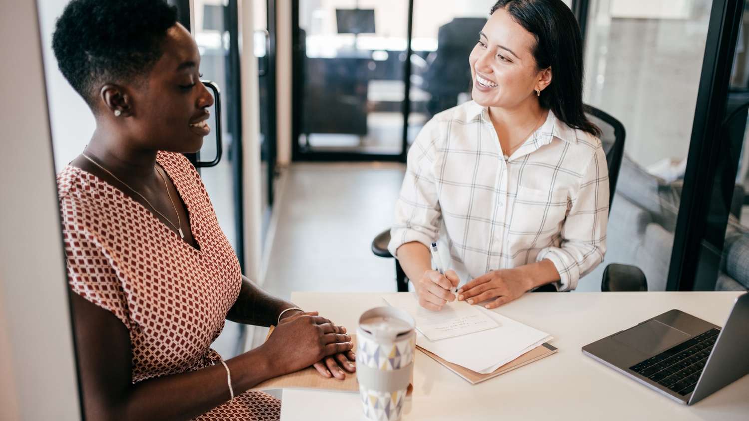Two women during a business meeting, one taking notes as the other looks on smiling.