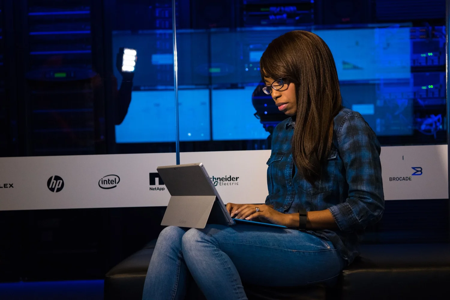 Woman in glasses and a plaid shirt types on a tablet in front of server racks and company logos.