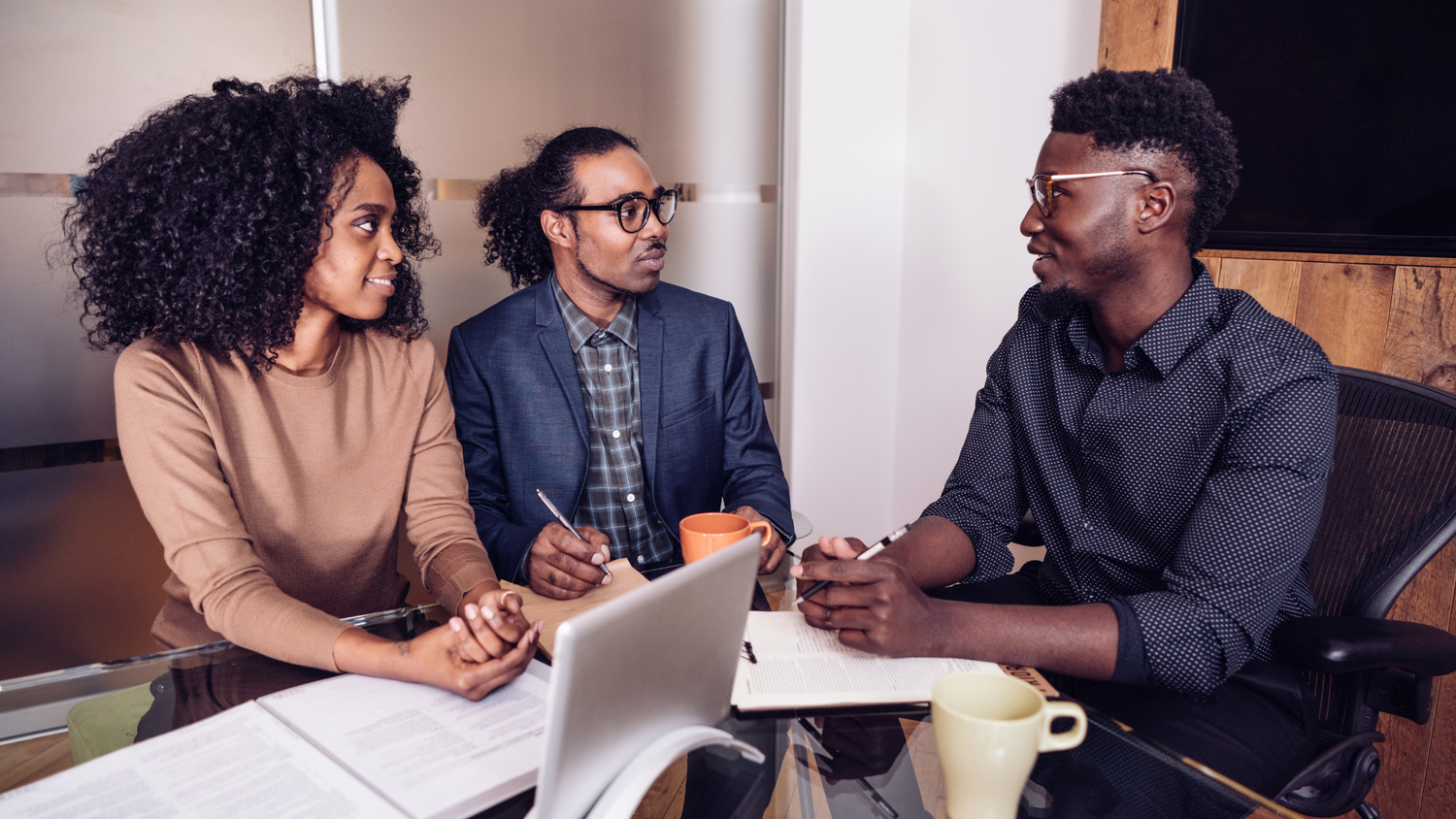 Three Black colleagues in business attire collaborate around a table, discussing notes and using a laptop.