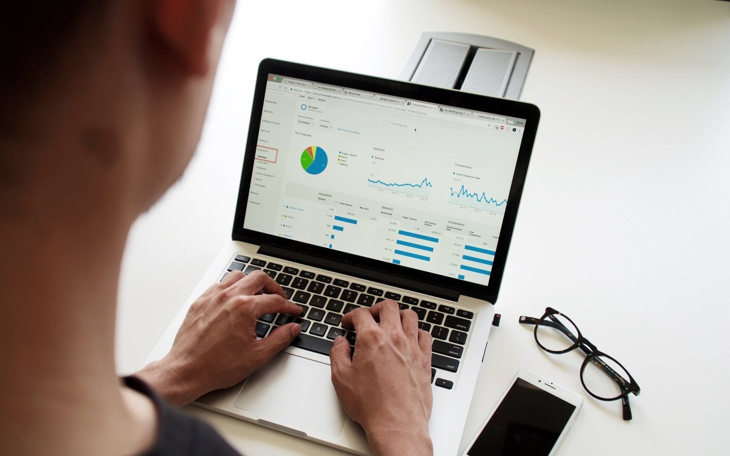 Person using a laptop displaying a Google Analytics dashboard with charts and graphs, glasses and a smartphone nearby.
