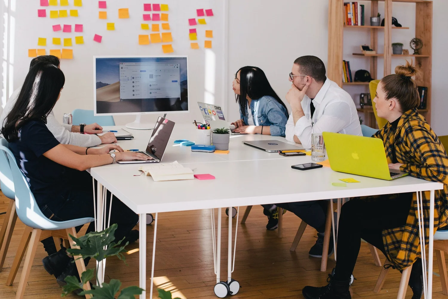 A diverse group of five people sit around a white table, focused on laptops and a large monitor displaying project management software.