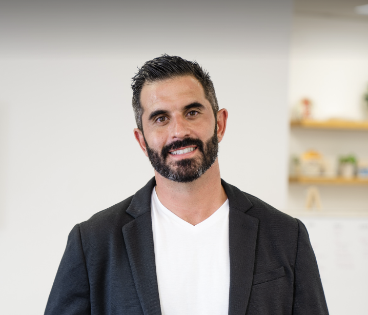 A man with dark hair, a beard, and a v-neck shirt under a dark blazer smiles at the camera.