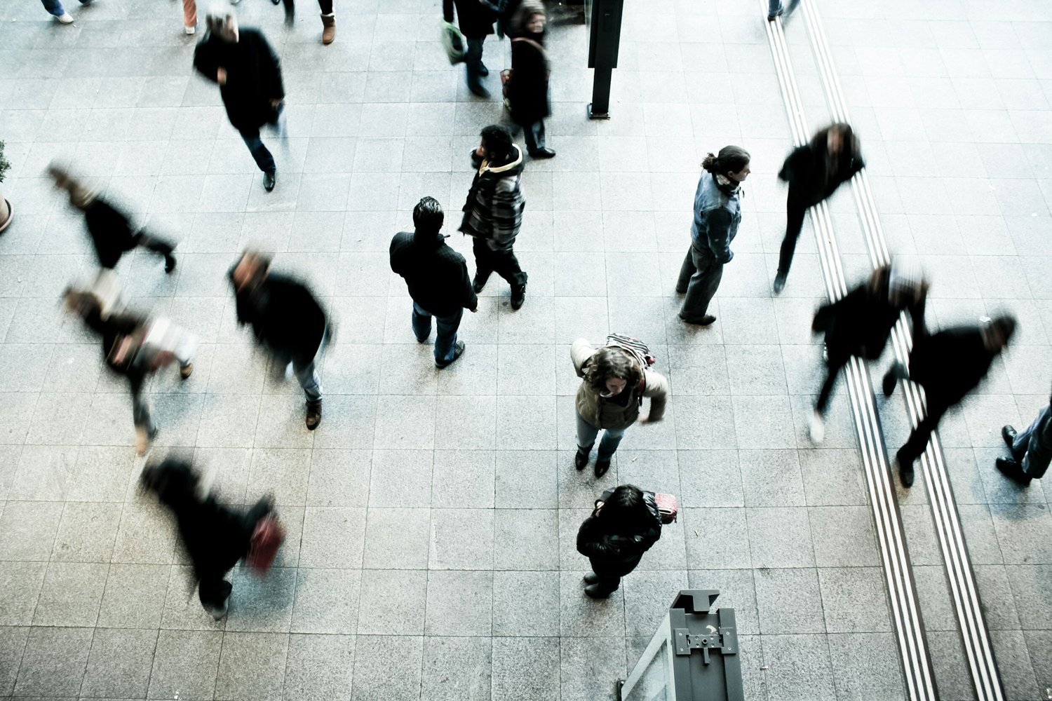 Blurry figures of people walking across a tiled floor, seen from above.