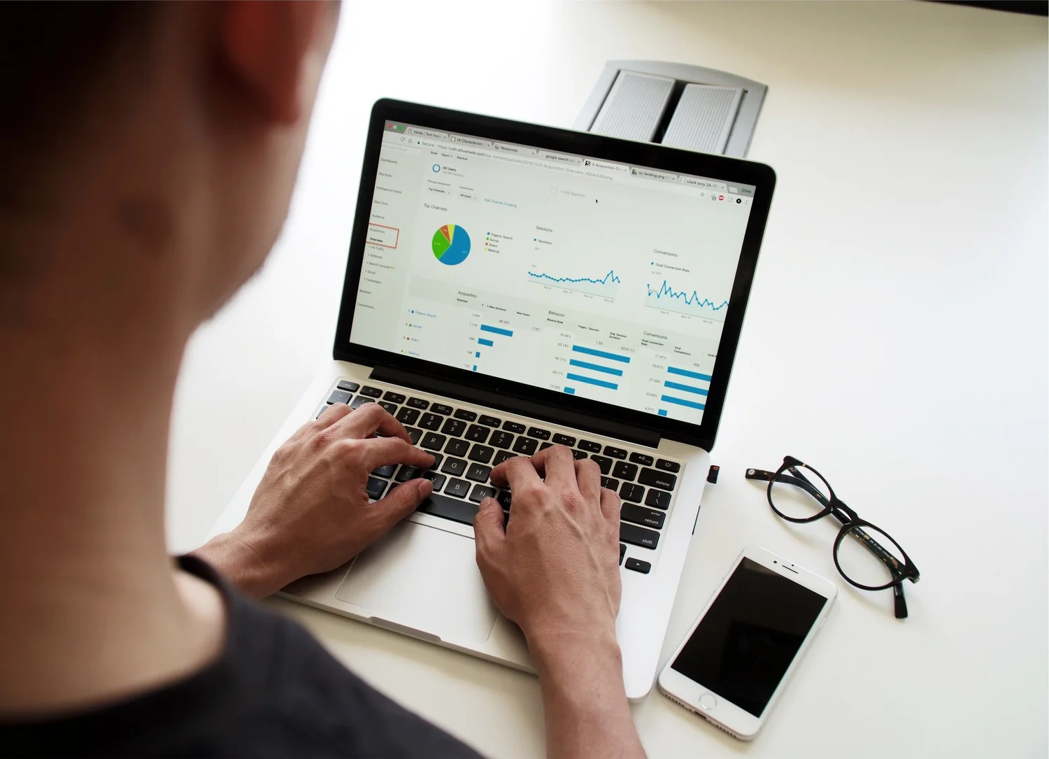 Person analyzing website data on a laptop screen showing charts and graphs, a smartphone and glasses on the desk.