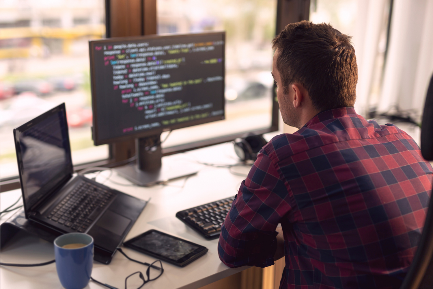 Developer in a plaid shirt sits at a desk looking at a computer screen with code; a laptop, coffee, and glasses are on the desk.