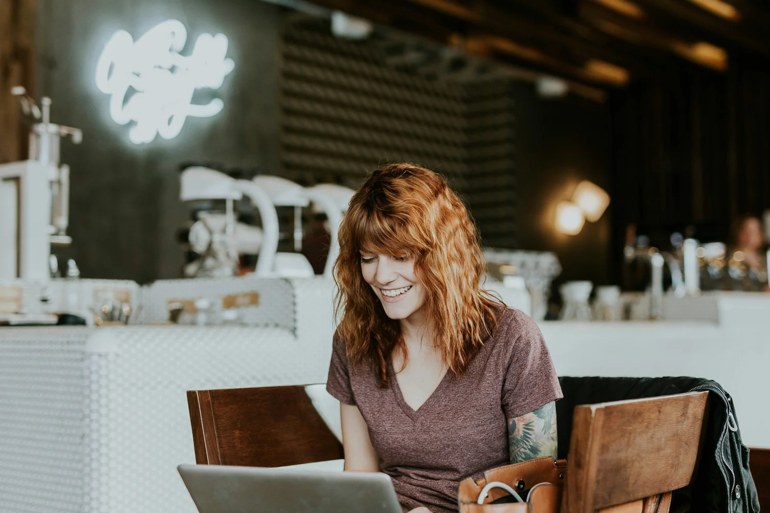 Woman with fiery red hair smiles while working on a laptop in a coffee shop, a neon sign in the background.