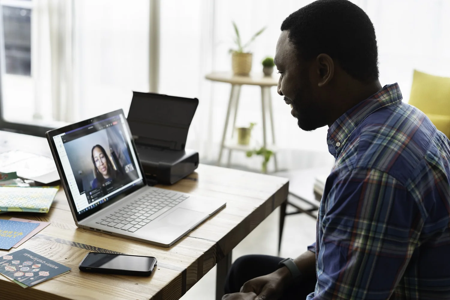 A Black man in a blue plaid shirt smiles while video conferencing with a woman on his laptop.
