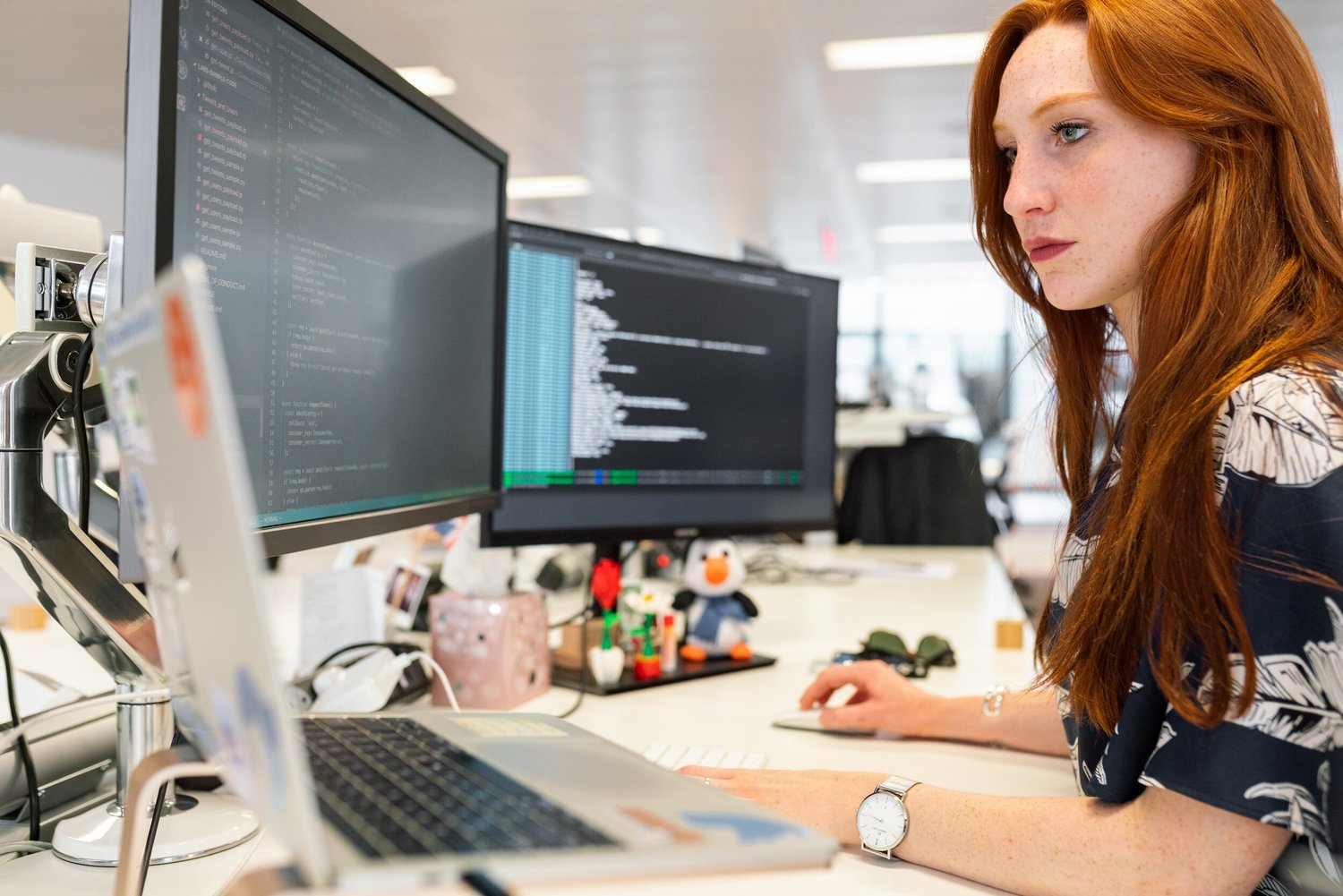 A woman with bright red hair sits at a desk focused on two screens displaying code, a laptop open in front of her.